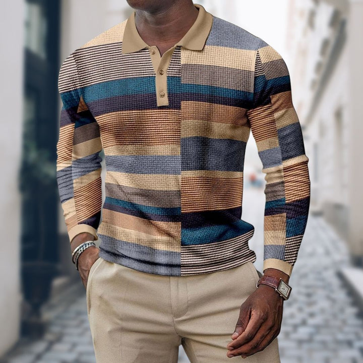 Mann trägt gestreiften Pullover in Beige, Blau und Braun, kombiniert mit beigen Hosen. Modischer Look auf gepflasterter Straße.