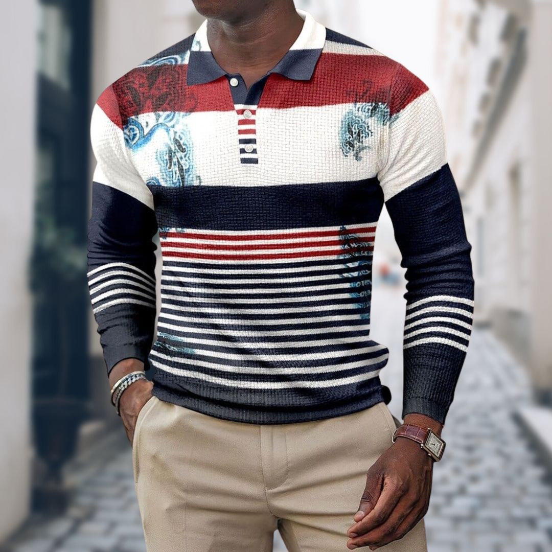 Mann trägt gestreiften Pullover in Rot, Blau und Weiß mit grafischen Mustern, kombiniert mit beigen Hosen. Modischer Herren-Look auf Stadtstraße.