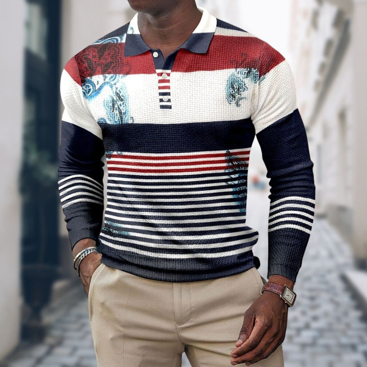Mann trägt gestreiften Pullover in Rot, Blau und Weiß mit grafischen Mustern, kombiniert mit beigen Hosen. Modischer Herren-Look auf Stadtstraße.