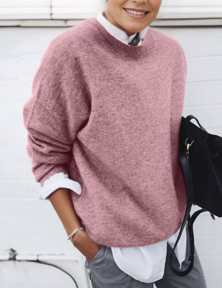 Frau in rosa Strickpullover, weißer Bluse und grauer Hose, hält schwarze Tasche. Modetrend Herbst, lässiger Stil, Damenmode.