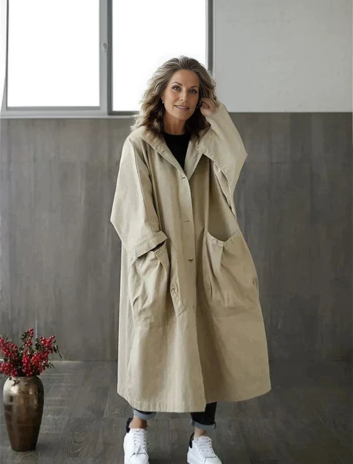 Frau in beigem Oversize-Mantel mit Kapuze, steht in modernem Raum. Modetrend, lässiger Stil, Herbstmode, bequeme Kleidung, Damenbekleidung.