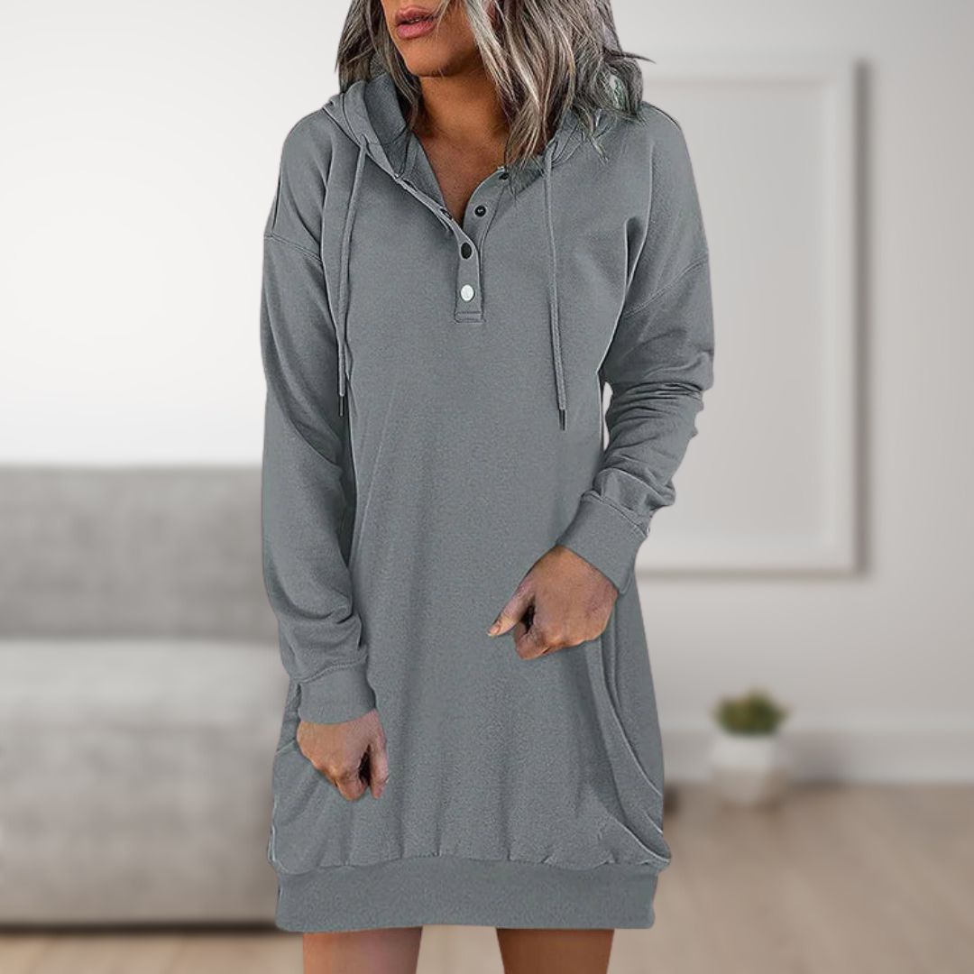 Frau trägt graues Kapuzen-Sweatshirt-Kleid mit Knopfleiste, lässig und bequem, ideal für Freizeitmode. Hintergrund unscharf, modernes Wohnzimmer.