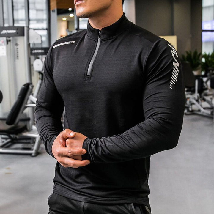 Mann im Fitnessstudio trägt schwarzes, langärmliges Sportshirt, muskulös, moderne Fitnessausrüstung im Hintergrund, Sportbekleidung, Training.