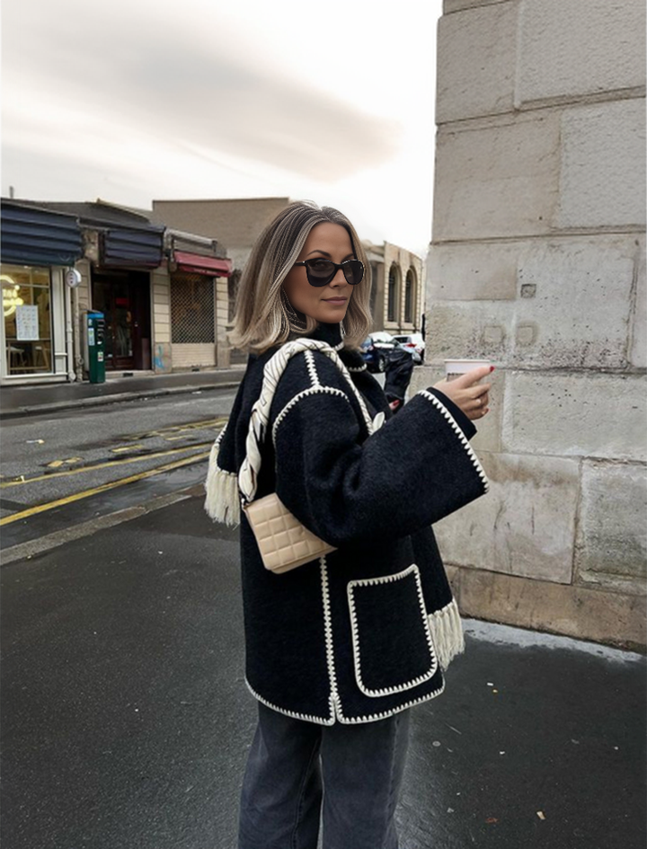 Frau mit schwarzem Mantel und Sonnenbrille hält Kaffeebecher, steht auf städtischer Straße. Modetrend, Herbstmode, Streetstyle.