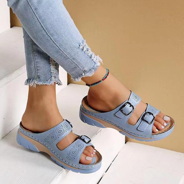 Damenfüße in blauen Sandalen mit Schnallen und Lochmuster, kombiniert mit zerrissenen Jeans. Modische Sommerschuhe auf weißer Treppe.
