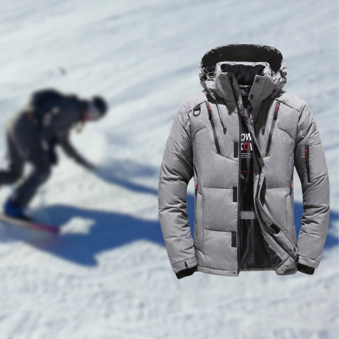 Graue Skijacke mit Kapuze im Vordergrund, ideal für Wintersport. Im Hintergrund unscharfer Skifahrer auf schneebedeckter Piste. Perfekt für Skiurlaub.