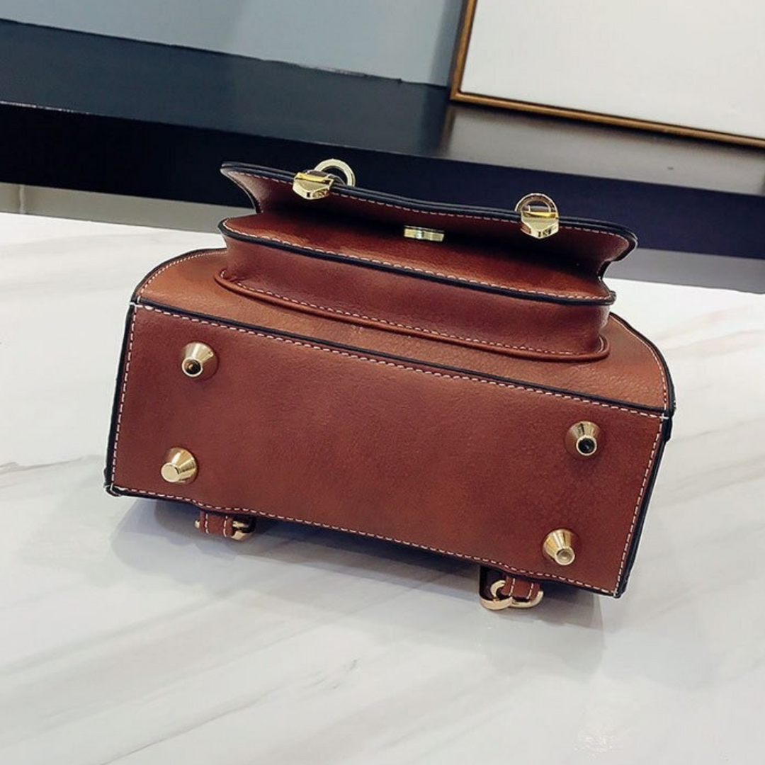 Luxuriöse braune Ledertasche mit goldenen Nieten und Verschlüssen auf weißem Marmortisch. Elegantes Design, hochwertige Verarbeitung, modisches Accessoire.