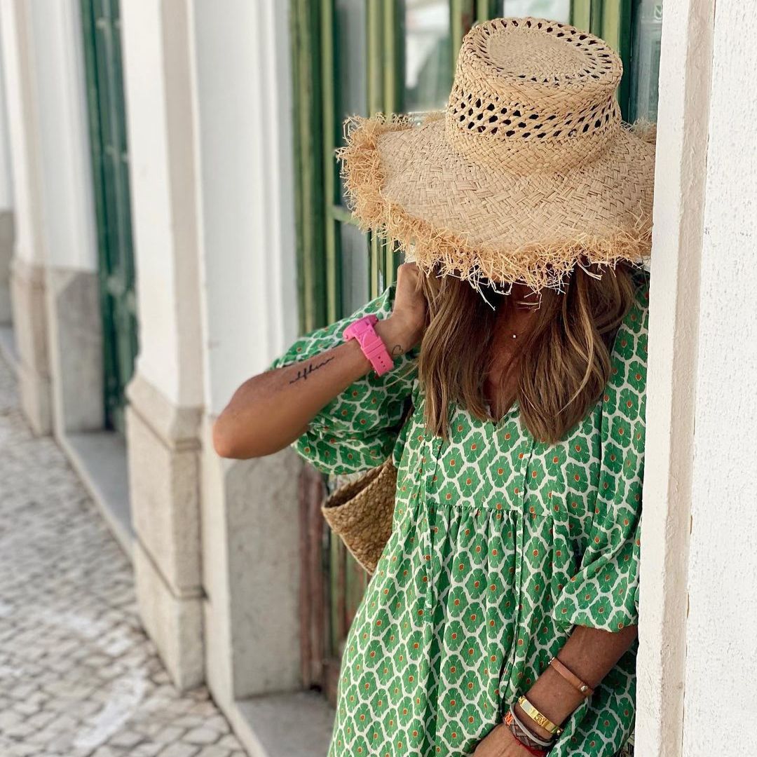 Frau mit grünem Kleid und Strohhut lehnt an weißer Wand. Sommermode, Boho-Stil, lässiger Look. Perfekt für sonnige Tage und Strandspaziergänge.