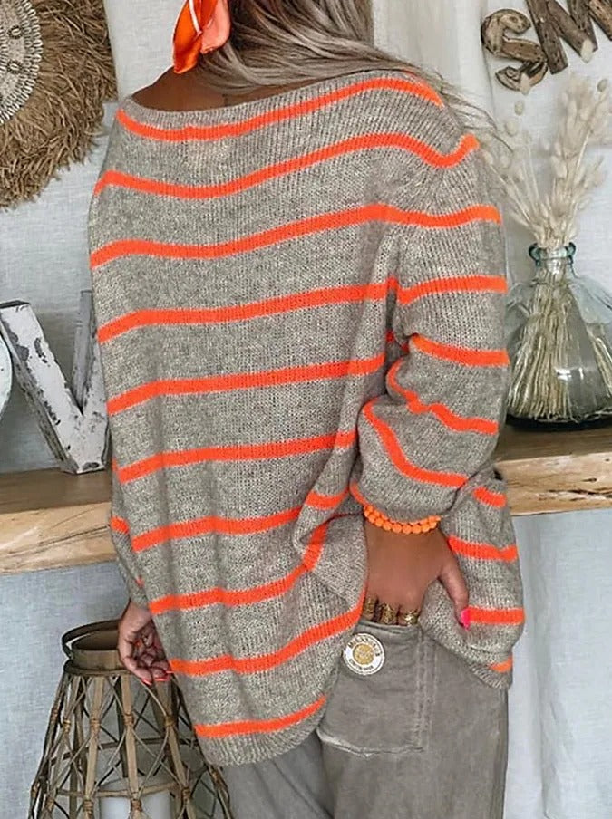 Frau in gestreiftem Pullover mit orangefarbenen Streifen, lässig gekleidet, steht vor rustikaler Dekoration. Herbstmode, Strickpullover, gemütlicher Stil.