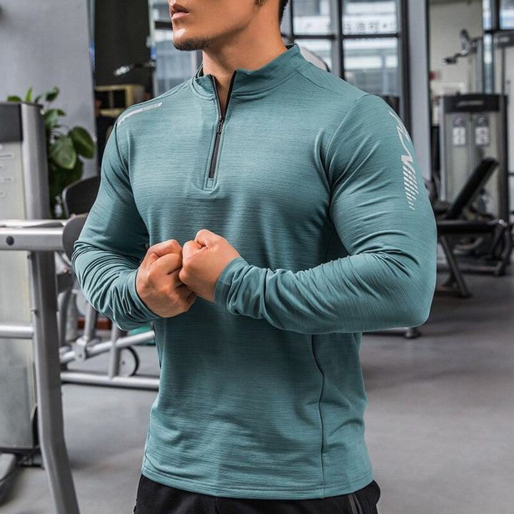 Mann im Fitnessstudio trägt grünes Langarm-Trainingsshirt, zeigt muskulöse Arme. Fitnessmode, Sportbekleidung, Männertraining, Gym-Outfit.