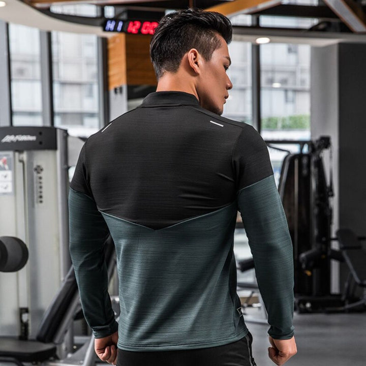 Mann im Fitnessstudio, trägt schwarzes und graues Sportshirt, steht vor Fitnessgeräten. Fitness, Sportbekleidung, Training, Gym.