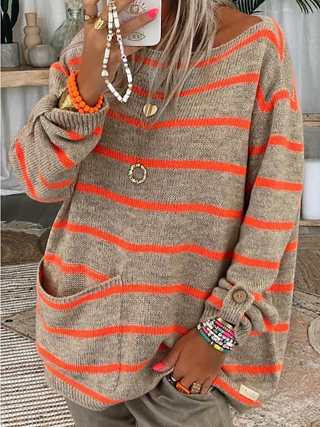 Frau in gestreiftem Oversize-Pullover mit orangefarbenen Streifen, hält Smartphone, trägt bunte Armbänder und Halsketten. Modetrend Herbst 2023.
