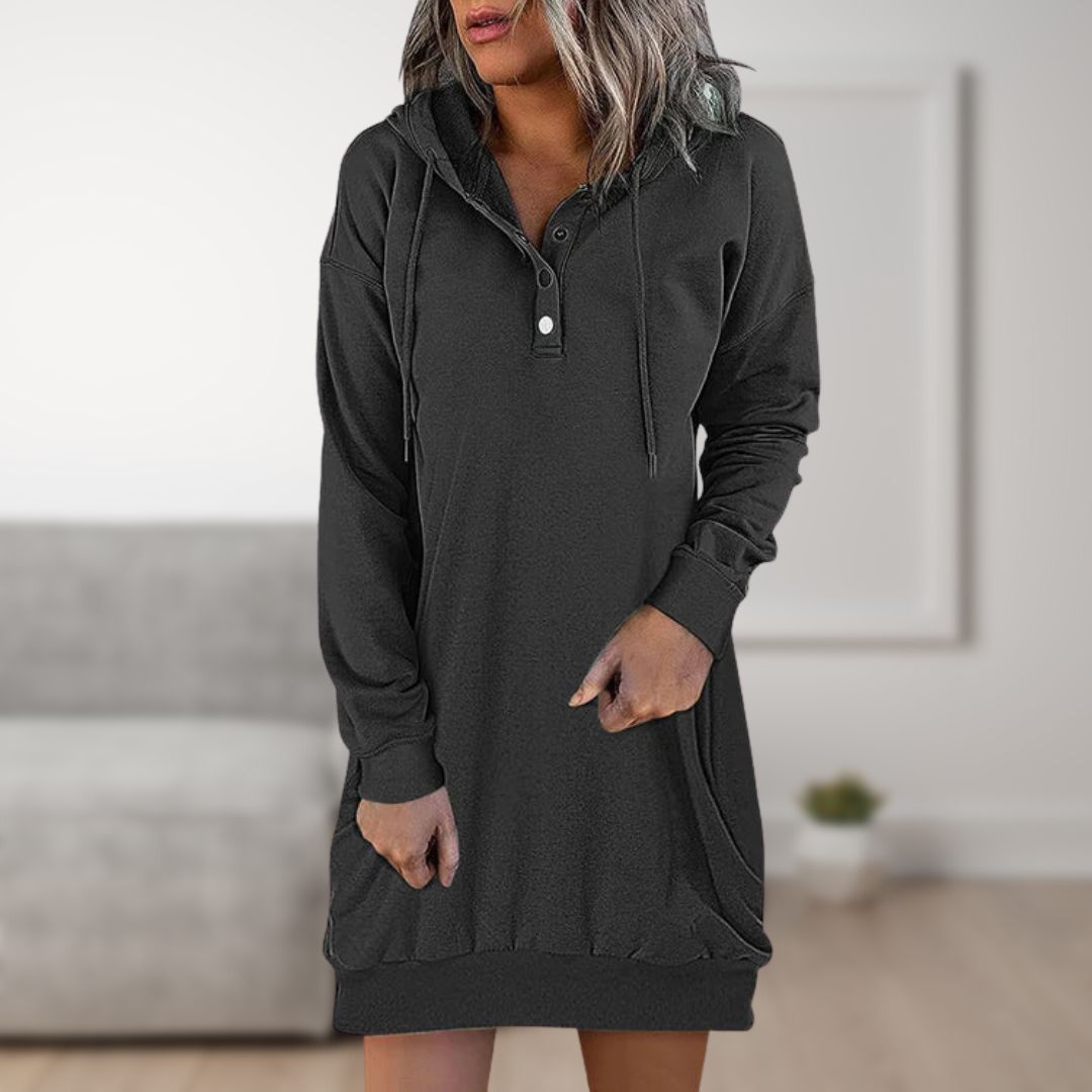 Frau in schwarzem Kapuzenkleid mit Knopfleiste, lässiger Stil, modernes Wohnzimmer im Hintergrund. Bequemes Freizeitoutfit für Damen.