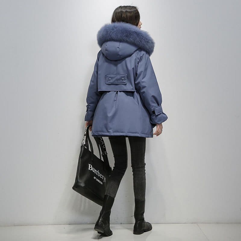 Frau in blauer Winterjacke mit Fellkapuze, schwarze Jeans und Stiefel, hält eine schwarze Einkaufstasche. Wintermode, Damenbekleidung, stilvoll.