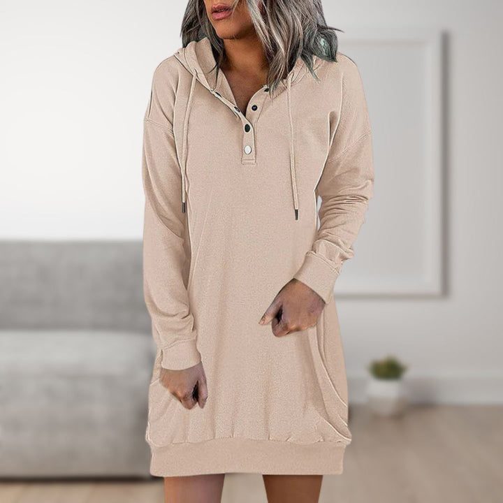 Frau in beigem Kapuzenkleid mit Knopfleiste, lässiger Stil, modernes Wohnzimmer im Hintergrund. Bequemes Freizeitoutfit, modische Damenbekleidung.