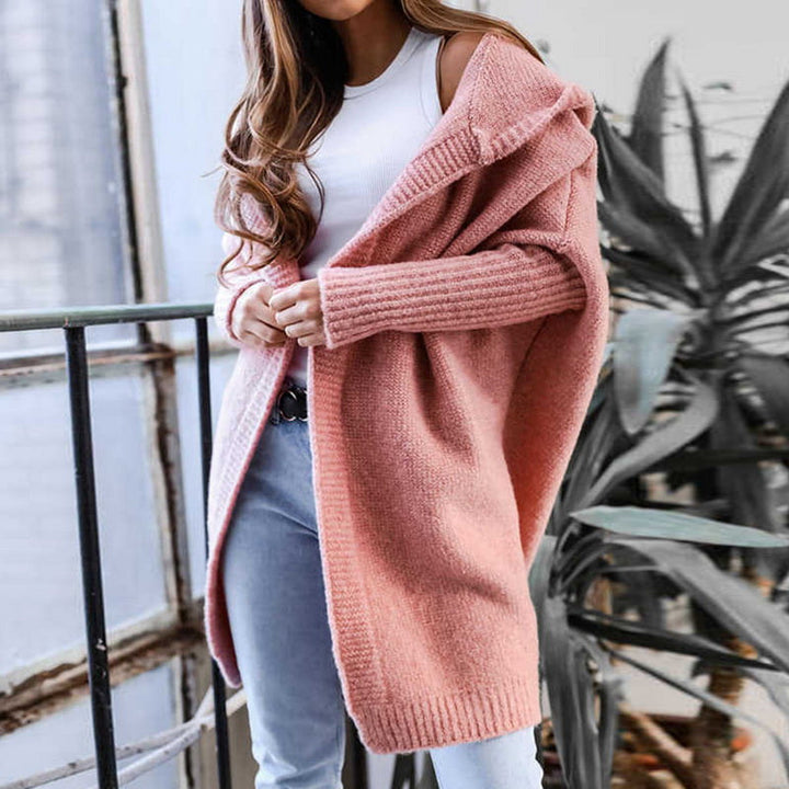 Frau in rosa Strickjacke und weißem Top, steht neben einem Fenster mit Pflanzen im Hintergrund. Modetrend, gemütlicher Look, Herbstmode.
