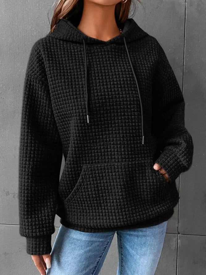 Frau trägt schwarzen, strukturierten Kapuzenpullover mit Kängurutasche und blauen Jeans vor grauem Hintergrund. Bequeme Freizeitmode.