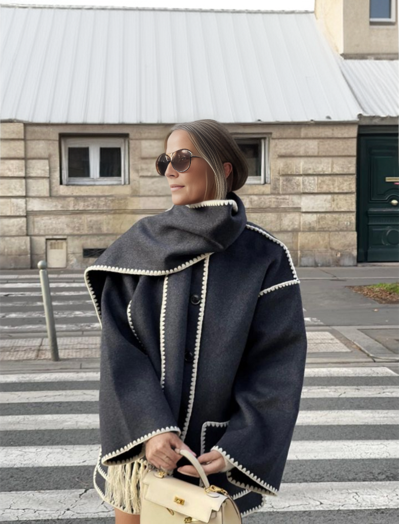 Frau in modischem, dunkelgrauem Mantel mit weißem Saum, Sonnenbrille und Handtasche auf Zebrastreifen vor städtischer Kulisse.