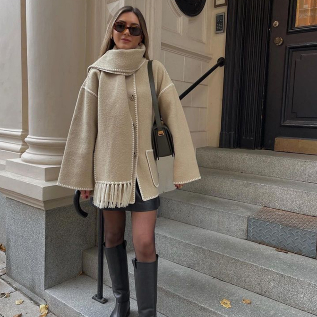 Frau in beigem Mantel und Schal, schwarze Stiefel, steht auf Treppe vor Stadthaus. Herbstmode, eleganter Look, urbane Kulisse.