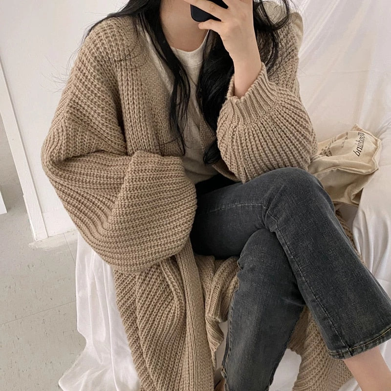 Frau in beigem Strickcardigan und Jeans sitzt auf einem weißen Sofa, hält ein Smartphone. Modetrend, gemütlicher Herbstlook, lässiger Stil.