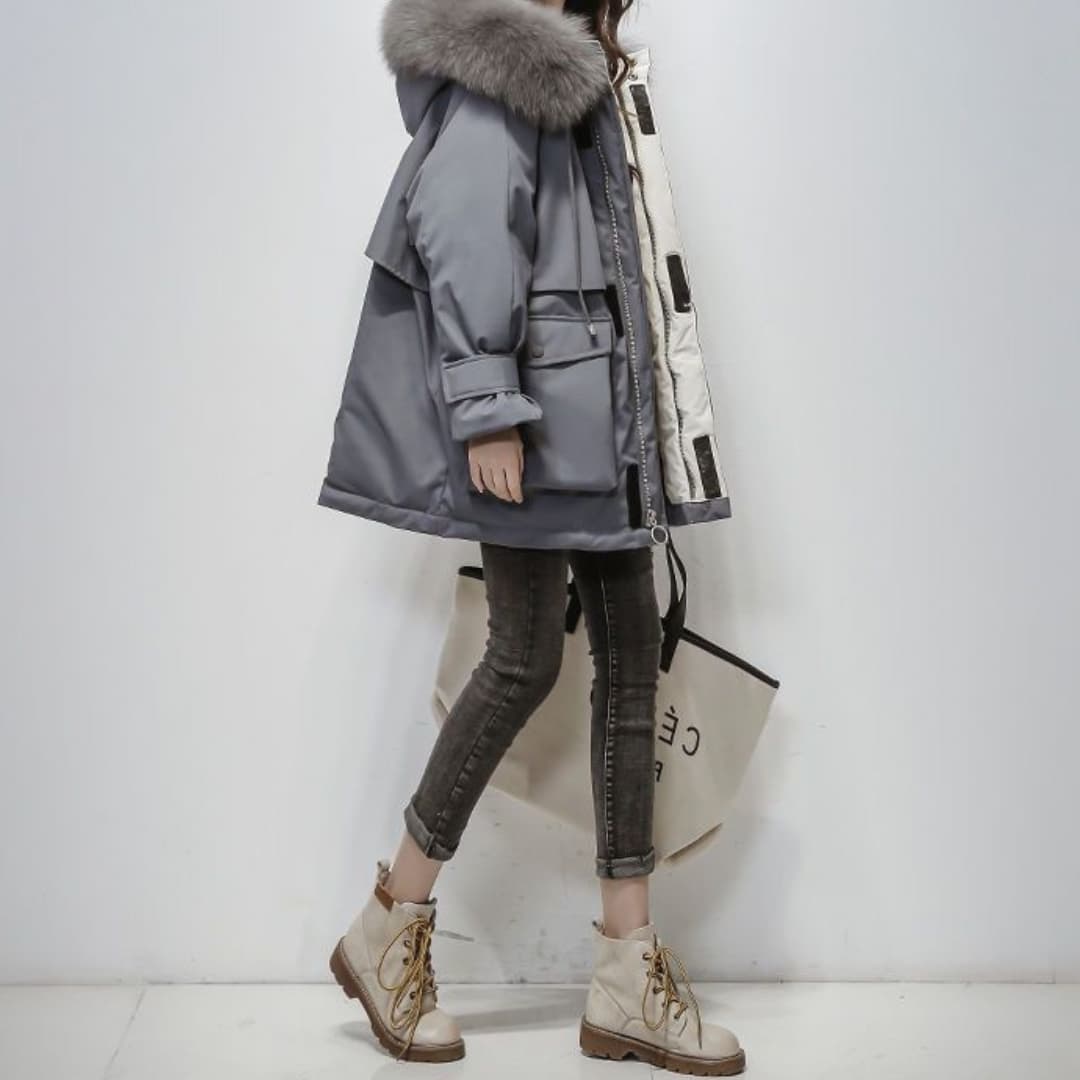 Dame in grauem Wintermantel mit Fellkapuze, trägt schwarze Jeans und beige Stiefel, hält eine Einkaufstasche. Modetrend, Wintermode, Damenbekleidung.