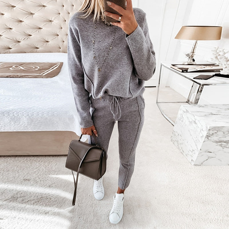 Frau in grauem Jogginganzug mit Handtasche im Schlafzimmer, moderne Einrichtung, gemütliches Ambiente, Mode, Freizeitkleidung, Innenraum.
