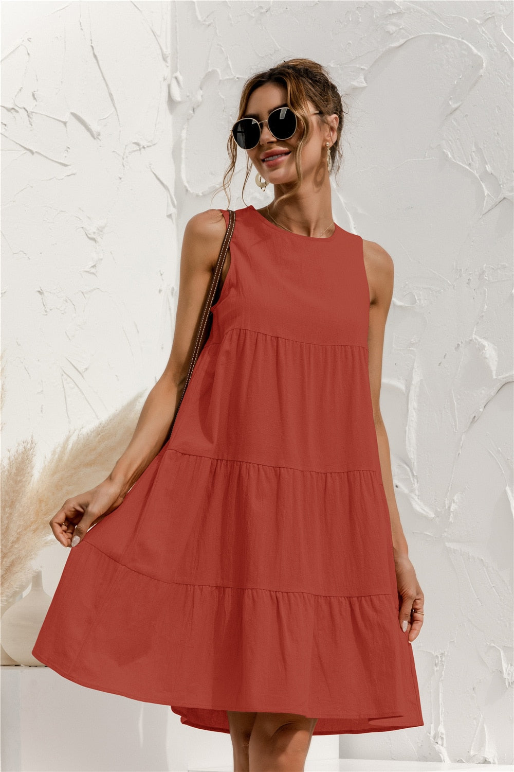 Frau in rotem Sommerkleid, lächelnd, mit Sonnenbrille vor weißer Wand. Modetrend, Damenmode, Sommeroutfit, lässiger Stil.