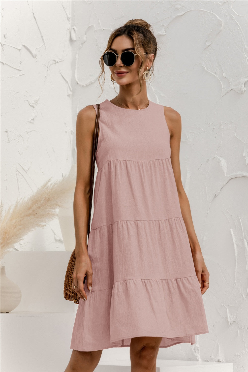 Frau in rosa Sommerkleid mit Sonnenbrille, brauner Tasche, vor weißer Wand. Modetrend, Damenmode, Sommeroutfit, lässiger Stil.