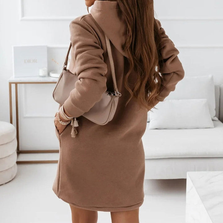 Frau in braunem Hoodie-Kleid mit beiger Handtasche, modernes Wohnzimmer, stilvolle Mode, gemütliches Interieur, elegantes Design.