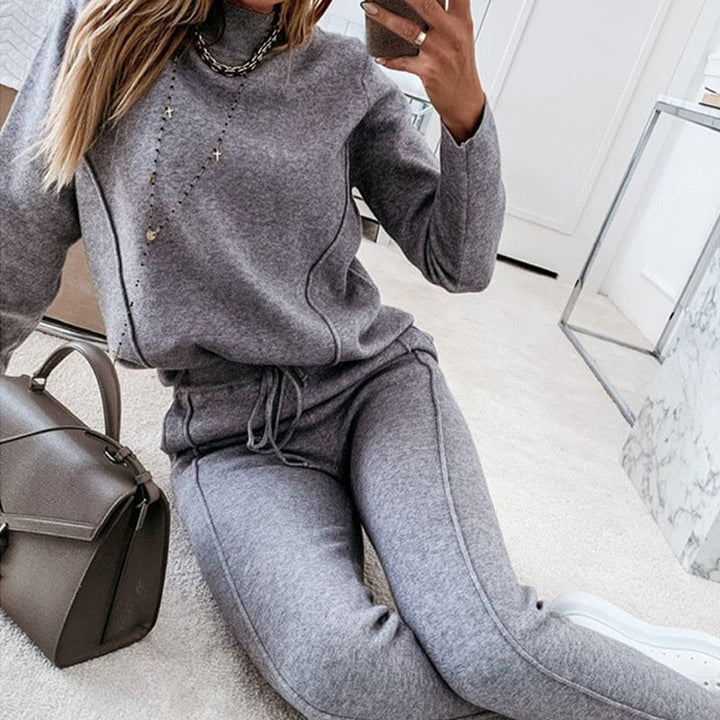Frau in grauem Jogginganzug macht Selfie, sitzt auf Teppichboden, mit brauner Handtasche und weißen Sneakers. Bequeme Freizeitmode, Indoor.
