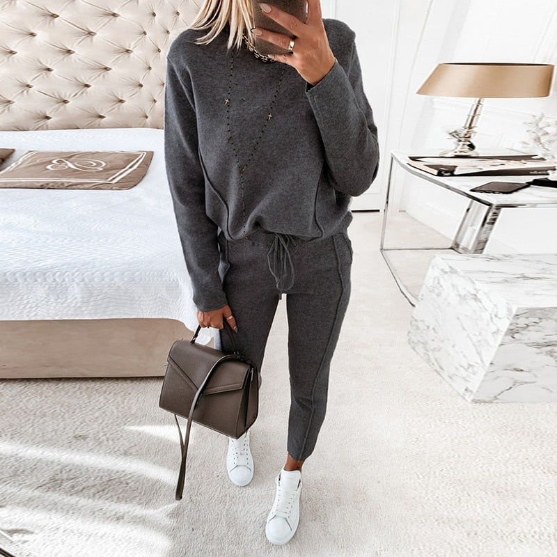 Frau in grauem Jogginganzug mit Handtasche und weißen Sneakers im Schlafzimmer. Moderne Einrichtung, Bett und Nachttisch im Hintergrund.