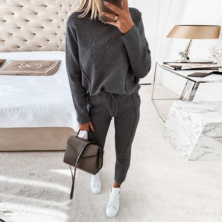 Frau in grauem Jogginganzug mit Handtasche und weißen Sneakers im Schlafzimmer. Moderne Einrichtung, Bett und Nachttisch im Hintergrund.