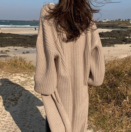 Frau in beigem Strickpullover am Strand, mit Blick aufs Meer. Herbstmode, Strickkleidung, Küstenlandschaft, entspannter Spaziergang.