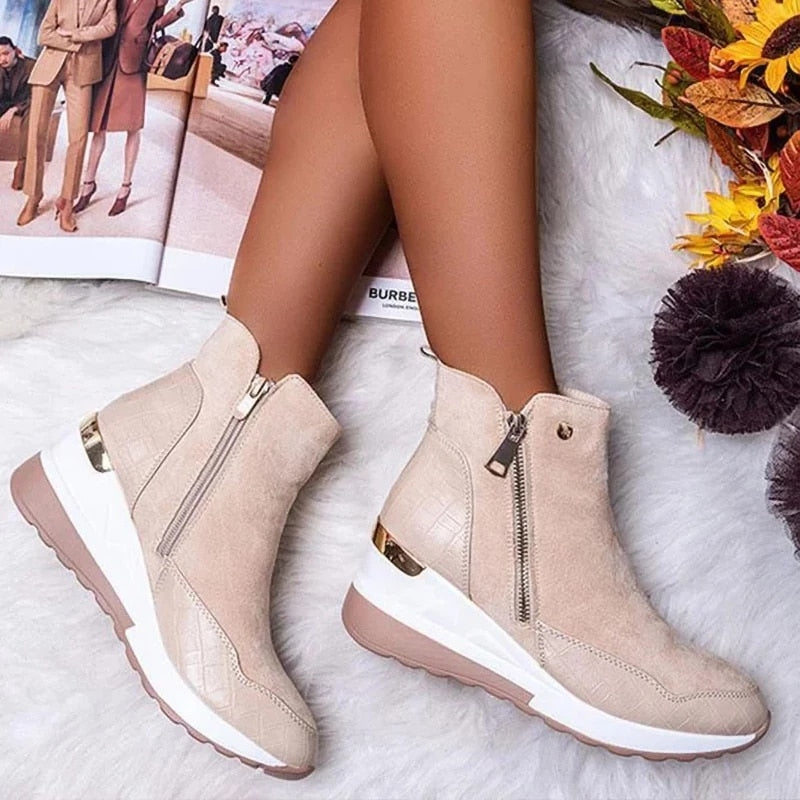 Beige Damenstiefeletten mit Keilabsatz und Reißverschluss auf weißem Teppich, umgeben von Herbstdekorationen und einer geöffneten Zeitschrift.
