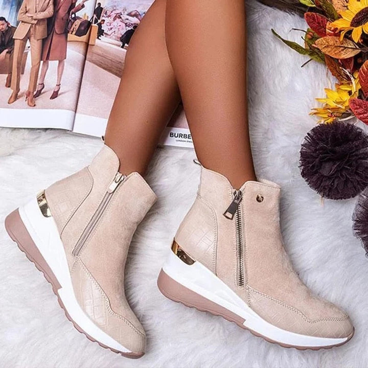 Beige Damenstiefeletten mit Keilabsatz und Reißverschluss auf weißem Teppich, umgeben von Herbstdekorationen und einer geöffneten Zeitschrift.