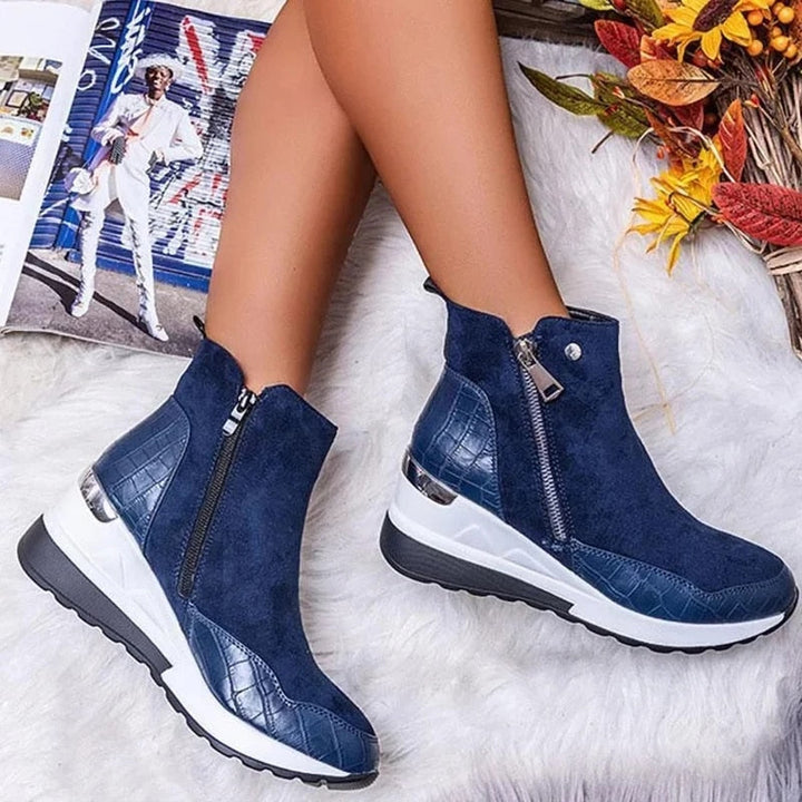 Blaue Damenstiefeletten mit Reißverschluss, kombiniert mit weißer Sohle und Krokodilleder-Details, auf Teppich mit Herbstdekorationen.