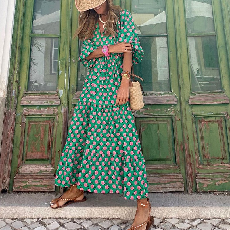 Frau in grünem Maxikleid mit rosa Blumenmuster, Strohhut und Sandalen vor grüner Holztür. Sommermode, Boho-Stil, lässiger Look.