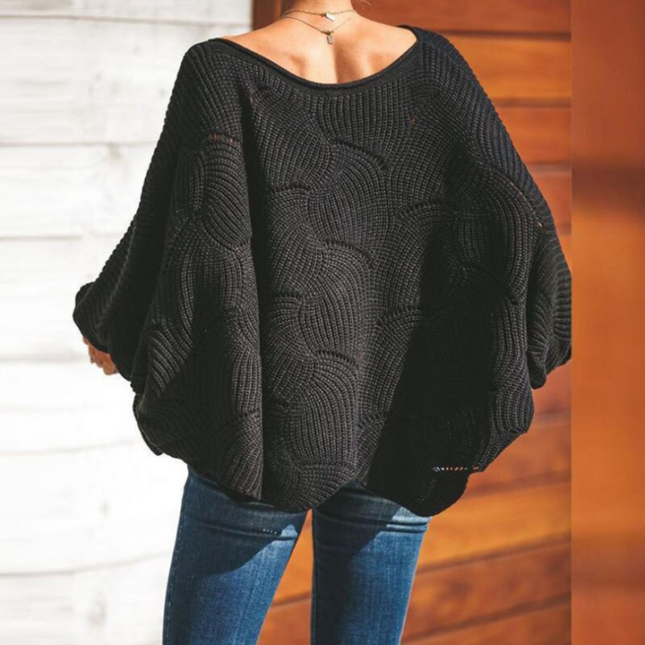 Schwarzer, gestrickter Oversize-Pullover mit Rundhalsausschnitt, getragen von einer Person in blauen Jeans, vor einer Holzwand.
