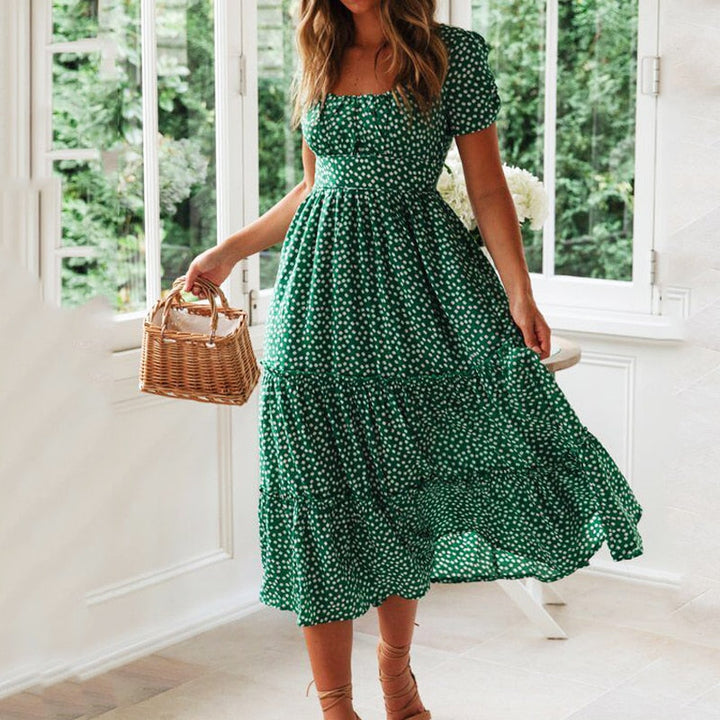 Frau in grünem Sommerkleid mit weißen Punkten, hält einen Korb in der Hand. Steht in einem hellen Raum mit großen Fenstern und Blick auf grüne Natur.