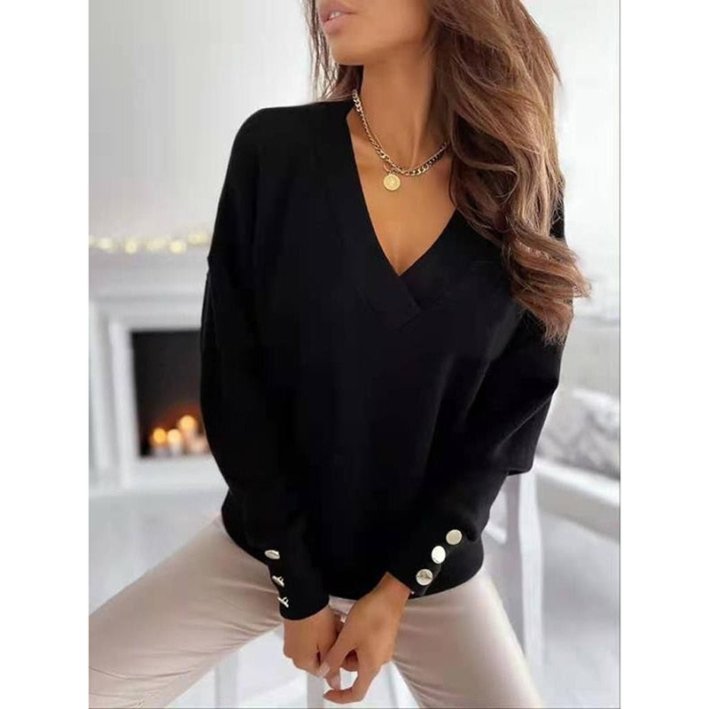 Frau in schwarzem V-Ausschnitt-Pullover mit Knöpfen, goldener Halskette, sitzt in modernem Wohnzimmer mit Kamin. Elegante Damenmode, Herbststil.