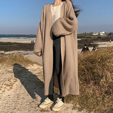 Frau am Strand in langem, beigem Strickcardigan, Jeans und Sneakers. Küstenlandschaft im Hintergrund. Modetrend, Strickmode, Strandspaziergang.