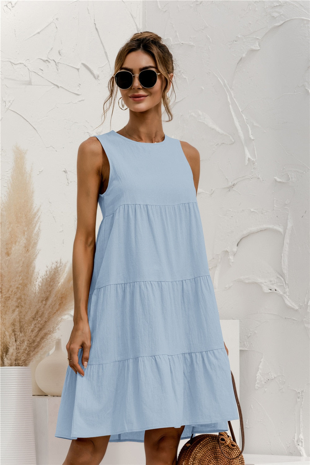Frau in hellblauem Sommerkleid, Sonnenbrille, mit Strohtasche vor weißer Wand. Modetrend, Sommermode, Damenkleid, lässiger Stil.