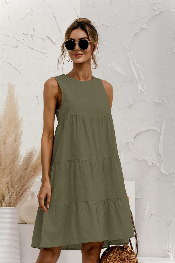 Frau in olivgrünem Sommerkleid, Sonnenbrille, mit Strohtasche vor weißer Wand. Elegante Damenmode, Sommeroutfit, Freizeitkleidung.