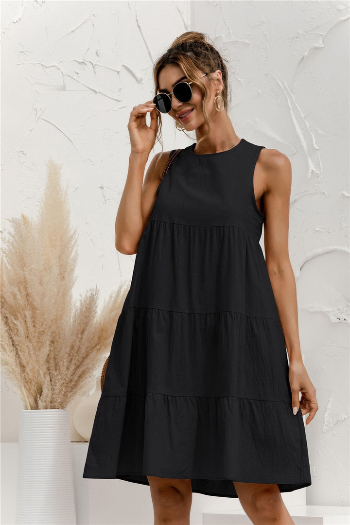 Frau in schwarzem Sommerkleid mit Sonnenbrille, lächelnd vor weißer Wand mit Pampasgras. Elegantes, ärmelloses Design, ideal für Sommermode.
