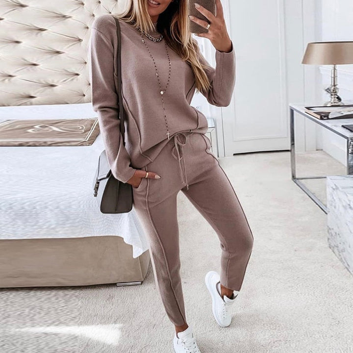 Frau in beigem Lounge-Set im Schlafzimmer, modisches Freizeitoutfit, gemütlicher Zweiteiler, weiße Sneaker, moderne Inneneinrichtung.