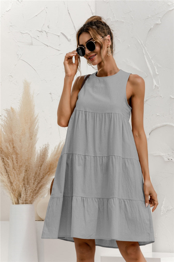 Frau in grauem Sommerkleid mit Sonnenbrille, vor weißer Wand mit Pampasgras. Modetrend, Damenmode, Sommeroutfit, lässiger Stil.