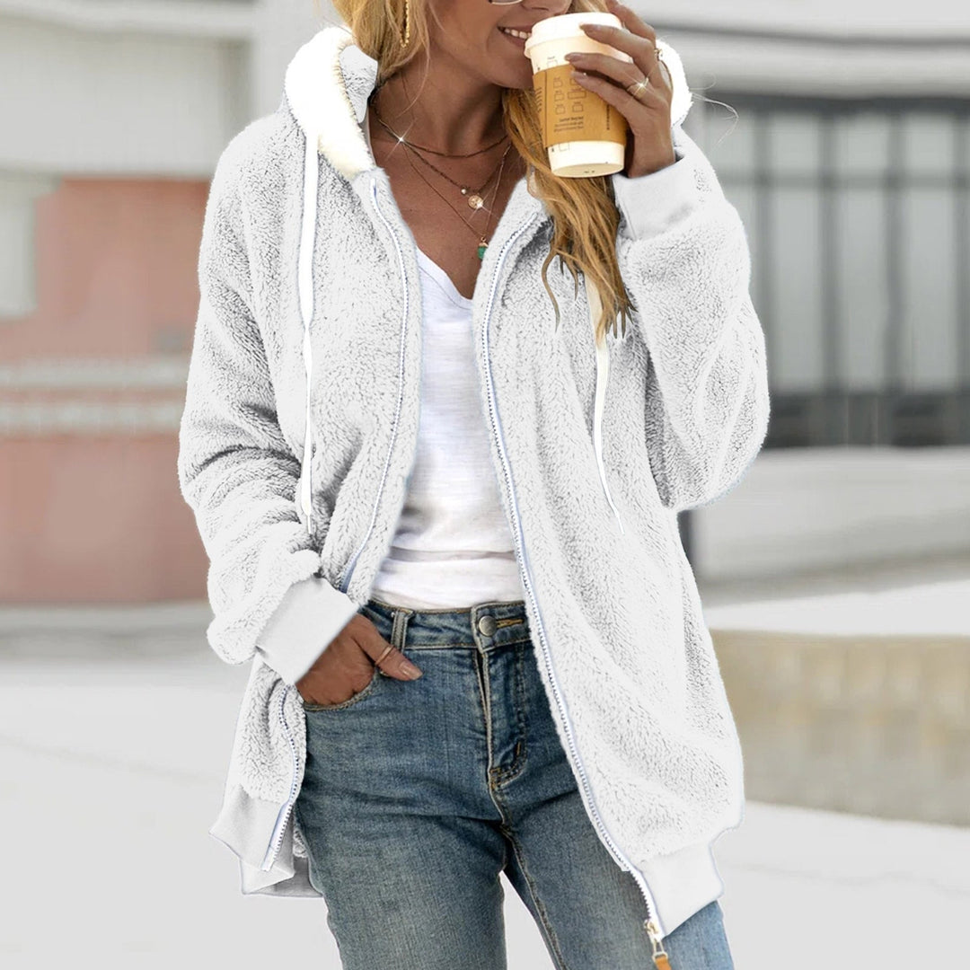 Frau in weißer Fleecejacke mit Kapuze hält Kaffeebecher, lässiger Look, Jeans, urbaner Hintergrund. Wintermode, gemütlicher Stil, Outdoor.