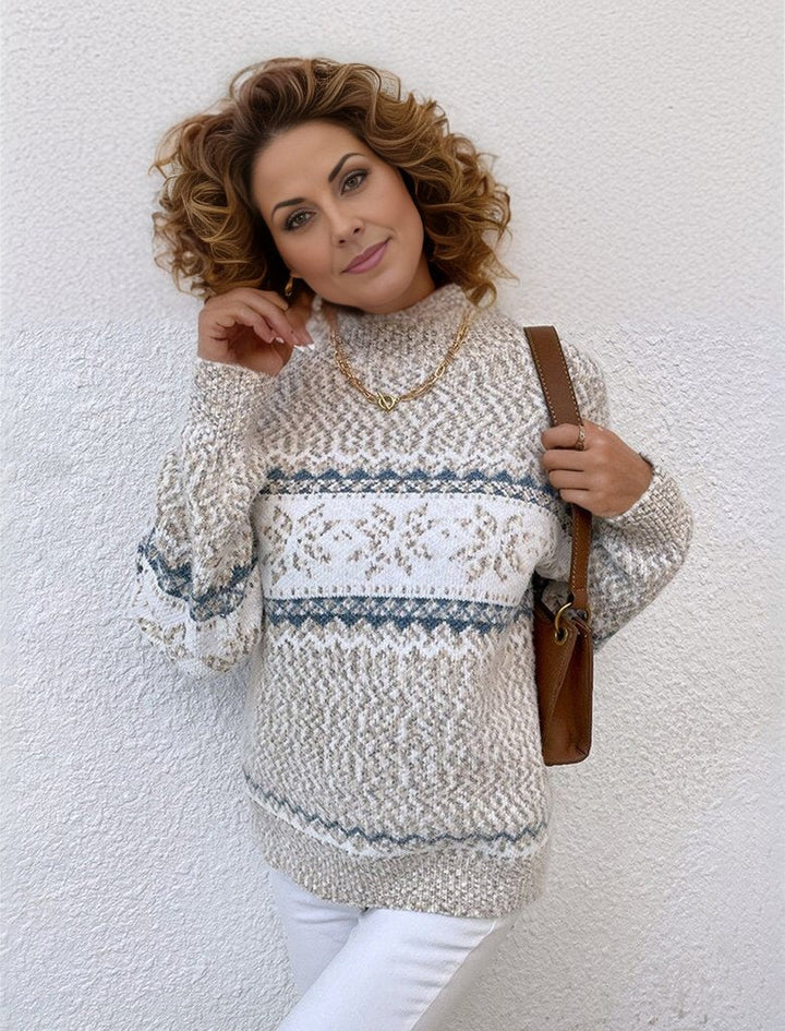 Eine Frau mit lockigem Haar trägt einen gestrickten Pullover mit blau-weißen Mustern und hält eine braune Tasche. Sie lehnt an einer weißen Wand.