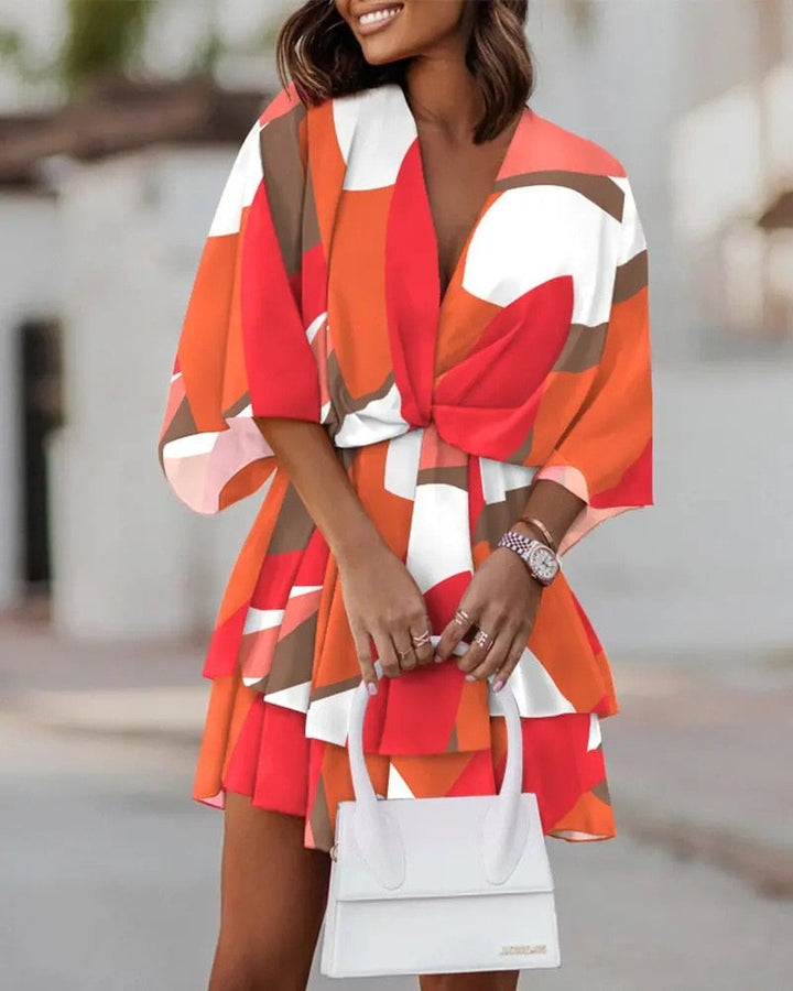 Frau in farbenfrohem, geometrisch gemustertem Kleid mit roten, weißen und braunen Tönen, hält weiße Handtasche. Sommermode, stilvoll, elegant.