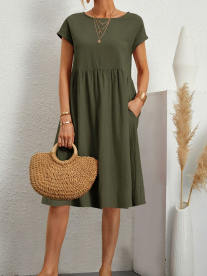 Frau in olivgrünem Sommerkleid mit kurzen Ärmeln, hält eine runde Strohtasche. Minimalistischer Stil, ideal für Freizeitmode und Sommertrends.